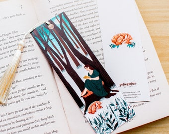 May include: A bookmark featuring a woman sitting on a tree stump in a forest reading a book. The bookmark has a blue, green, and white color scheme with a white tassel. The text on the bookmark reads "julia kestner designs".
