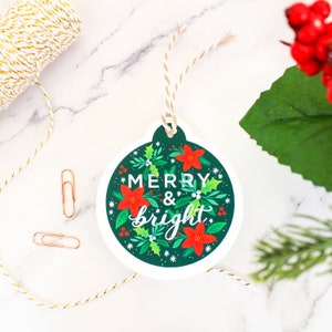 May include: A green and white Christmas ornament gift tag with red poinsettia flowers and holly leaves. The tag has the text "Merry & bright" in white lettering.