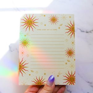May include: A cream-colored notepad with gold lined paper and a gold and red sun design. The notepad has the text "ONE DAY AT A TIME" printed on the front.
