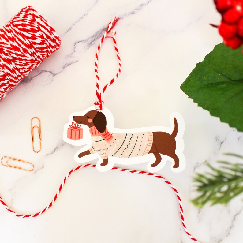 May include: A white gift tag shaped like a dachshund dog wearing a pink and green sweater and carrying a red gift box. The tag is attached to a red and white twine.
