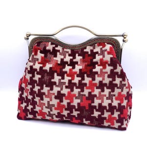 Może przedstawiać: Wzorzysta torebka z metalowym zapięciem i uchwytem. Torba ma wzór pepitki w odcieniach czerwieni, bordowego i kremowego. Metalowa rama ma wykończenie ze starego mosiądzu, dodając akcesorium charakteru vintage.