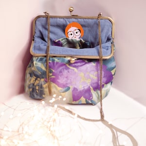 Könnte beinhalten: Eine Handtasche im Vintage-Stil mit einem Blumenmuster in Lila, Blau und Gelb. Die Tasche hat einen Messingverschluss und einen Kettenriemen. Das Innere ist mit blauem Stoff gefüttert. Eine Cartoonfigur mit roten Haaren und Brille schaut heraus.