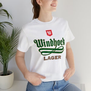 Puede incluir: Camiseta blanca con la inscripción "Windhoek Lager" en verde y marrón, con un logotipo rojo en forma de escudo encima. La camiseta de manga corta está hecha de un material suave.