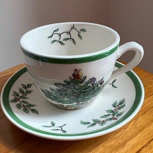May include: A white teacup and saucer with a green rim and a Christmas tree design. The tree is decorated with ornaments, a star, and a small figure of Santa Claus.