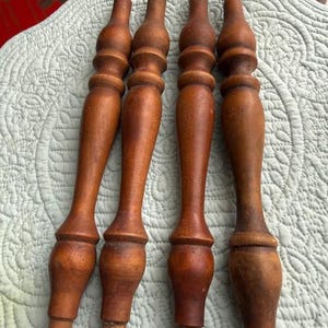 May include: Four antique wooden furniture legs with a warm brown finish. The legs have a turned design with a bulbous center and tapered ends. They are resting on a light blue quilted surface.