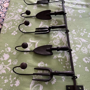 Pode incluir: Um suporte de ferramentas de jardim de metal preto com cinco ganchos. Cada gancho tem a forma de uma ferramenta de jardim diferente, incluindo uma pá, um ancinho e um garfo. O suporte está montado em uma parede.