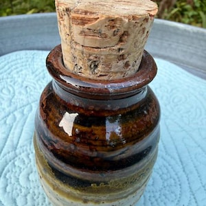 Pode incluir: Um pequeno pote de cerâmica cilíndrico com um esmalte brilhante de vários tons, passando do castanho escuro no topo para um bege claro salpicado na base. O pote é selado com uma rolha de cortiça natural espessa, texturizada e castanha.