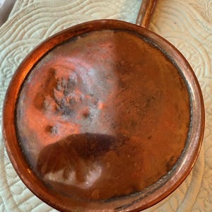 May include: A vintage copper pan with a long handle. The pan has a round, shallow bowl with a slightly raised rim. The copper has a warm, reddish-brown color with some darker areas, suggesting age and use. The handle is also copper.