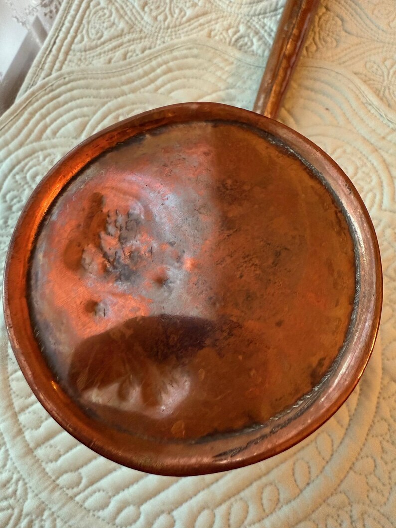 May include: A vintage copper pan with a long handle. The pan has a round, shallow bowl with a slightly raised rim. The copper has a warm, reddish-brown color with some darker areas, suggesting age and use. The handle is also copper.