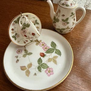 May include: A miniature tea set featuring a white teapot, teacup, and saucer. The set is decorated with a strawberry and leaf pattern, with gold trim. The teacup is suspended in a gold-colored metal holder, and the saucer is round.