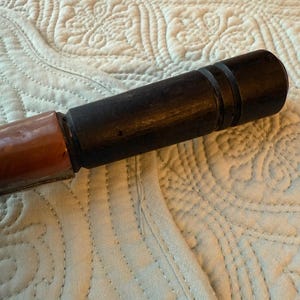 May include: A dark brown wooden object with a cylindrical shape, featuring a lighter brown section at one end. The object has a smooth, polished appearance and appears to be a handle or part of a larger tool.