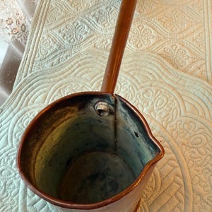 May include: A vintage copper pitcher with a long wooden handle. The pitcher has a spout and a rounded body. The handle is dark brown at the end. The copper has a warm, reddish-brown hue, with some areas showing a darker patina.