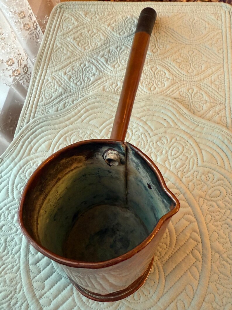 May include: A vintage copper pitcher with a long wooden handle. The pitcher has a spout and a rounded body. The handle is dark brown at the end. The copper has a warm, reddish-brown hue, with some areas showing a darker patina.