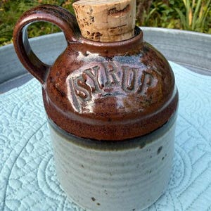 May include: A two-toned ceramic syrup jug with a glossy dark brown upper section and a matte light gray lower section. The word SYRUP is embossed in capital letters on the shoulder. It features a curved handle and a natural cork stopper.