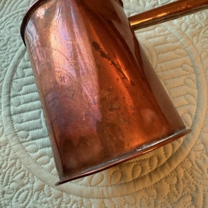 Puede incluir: Una jarra de cobre con un asa larga y un pico. La jarra tiene una superficie brillante y reflectante con algunas manchas más oscuras. El asa es de un color cobre similar. La jarra probablemente se utiliza para verter líquidos.