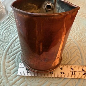May include: A copper-colored metal pitcher with a wooden handle. The pitcher has a spout and a cylindrical body. A ruler is placed next to the pitcher, showing measurements in inches.