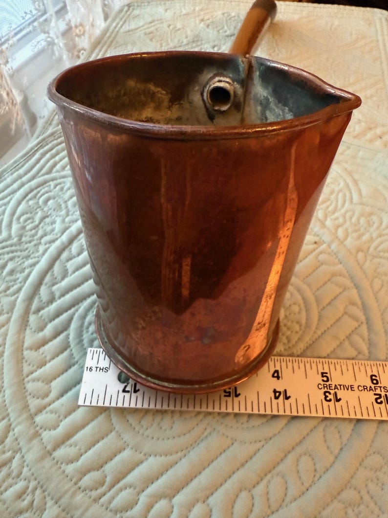 May include: A copper-colored metal pitcher with a wooden handle. The pitcher has a spout and a cylindrical body. A ruler is placed next to the pitcher, showing measurements in inches.