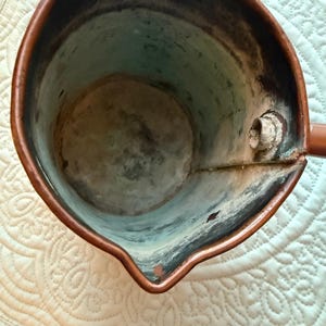 May include: A copper-colored, antique-style pitcher with a spout and handle. The interior shows signs of age with a patina of blue and green hues. The pitcher sits on a white, quilted surface.