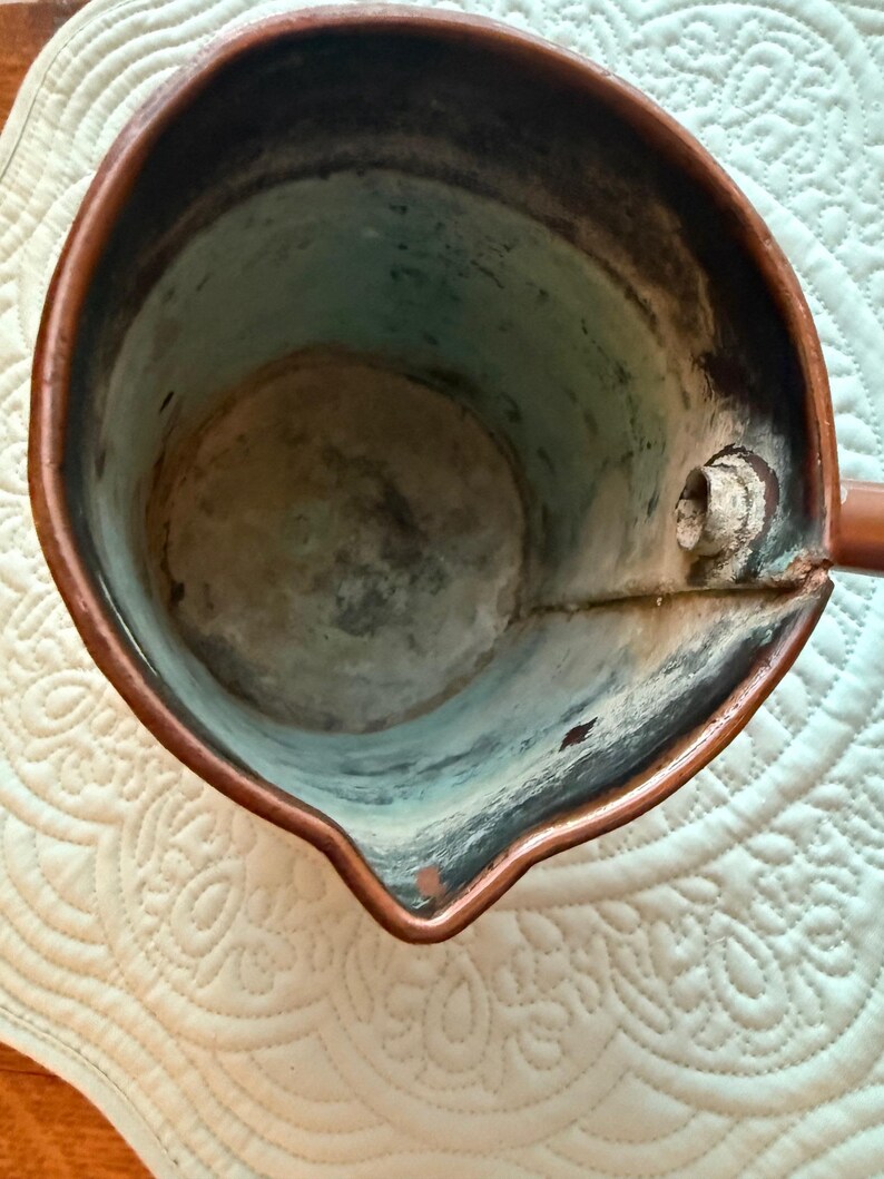 May include: A copper-colored, antique-style pitcher with a spout and handle. The interior shows signs of age with a patina of blue and green hues. The pitcher sits on a white, quilted surface.