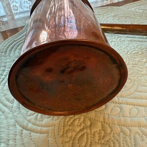 May include: A copper pitcher with a spout and handle. The pitcher has a rounded base and a polished, reflective surface. The copper has a warm, reddish-brown hue, and the pitcher appears to be vintage or antique.