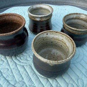 May include: A set of four small, round ceramic cups with a dark, mottled brown and black glaze on the exterior. The rims and interiors feature a lighter, speckled beige and cream finish, showcasing a hand-thrown pottery aesthetic with visible texture.