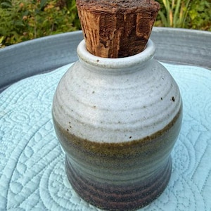 Pode incluir: Um frasco de cerâmica com corpo arredondado e esmalte de dois tons, que transita de um branco sujo salpicado no topo para um castanho escuro na base. O frasco é rematado com uma rolha de cortiça natural castanha, espessa e texturizada.