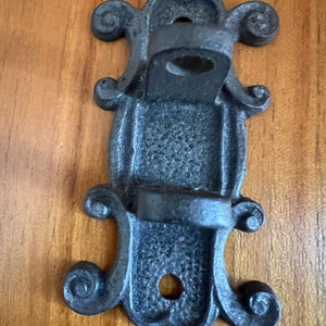 Könnte beinhalten: Schwarzer Wandhaken aus Metall mit dekorativem Schnörkeldesign. Der Haken hat zwei Aufhängepunkte.