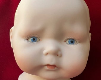 May include: A porcelain doll head with blue eyes and a painted expression. The head is mounted on a white stand.