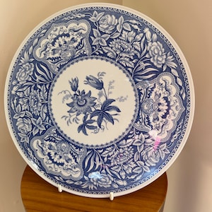 以下が含まれることがあります： 青と白のフローラルパターンが施された白いセラミックプレート。プレートは縁が高く、大きな開いた花を咲かせた花を中央にデザインしています。