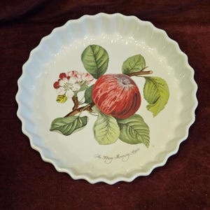 Könnte beinhalten: Eine weiße, gewellte Keramik-Tarteform mit einer zentralen botanischen Illustration eines roten Apfels mit grünen Blättern und weißen Blüten. Der Text The Early Morning Apple ist in schwarzer Schrift am unteren Rand aufgedruckt.