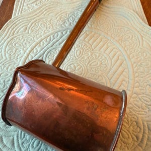 May include: An antique copper ladle with a long wooden handle and a dark brown grip. The copper has a rich, aged patina, and the ladle's shape suggests it was used for cooking or serving. The handle is attached to a rectangular bowl.