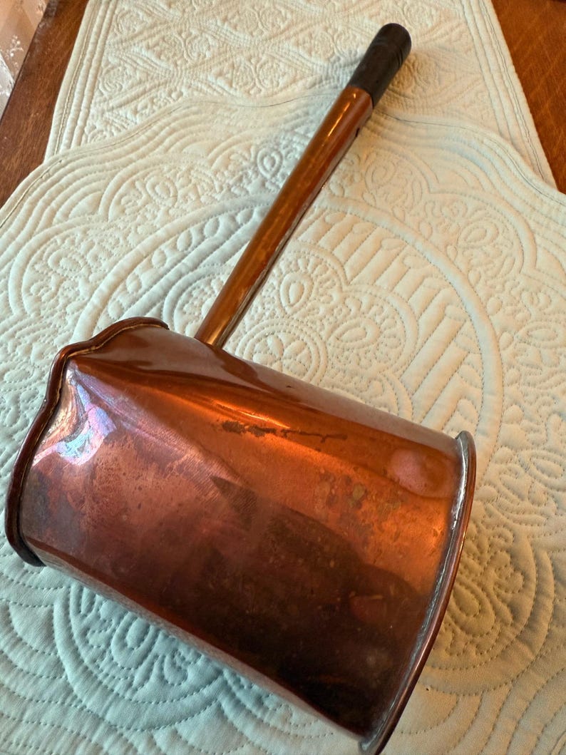 May include: An antique copper ladle with a long wooden handle and a dark brown grip. The copper has a rich, aged patina, and the ladle's shape suggests it was used for cooking or serving. The handle is attached to a rectangular bowl.