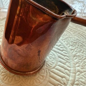 May include: A copper-colored, angled pitcher with a long handle. The pitcher has a shiny, reflective surface and a slightly flared rim. The handle is a darker shade of brown. The pitcher is likely vintage or antique.