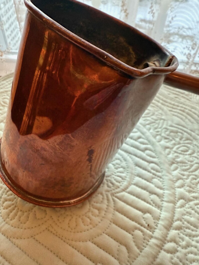 May include: A copper-colored, angled pitcher with a long handle. The pitcher has a shiny, reflective surface and a slightly flared rim. The handle is a darker shade of brown. The pitcher is likely vintage or antique.