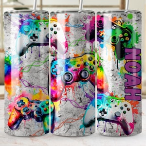 May include: A stainless steel tumbler with a design of colourful video game controllers on a textured grey background. The name "NOAH" is displayed vertically in purple. The design includes paint splatters and colourful accents.