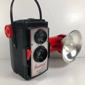 May include: A black and red vintage Imperial Deluxe Twin Lens Reflex camera with a built-in flash. The camera has a red carrying handle and a red flash attachment.