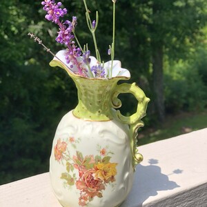 May include: A small, green and white porcelain pitcher with a floral design and a gold trim. The pitcher is filled with purple lavender flowers.