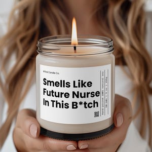 Puede incluir: Una vela blanca con una etiqueta negra que dice "#Viral Candle Co. Smells Like Future Nurse In This B*tch" y "Hand-poured 100% natural soy 50-60h | 19 oz | 265g". La vela está encendida y la llama es visible.