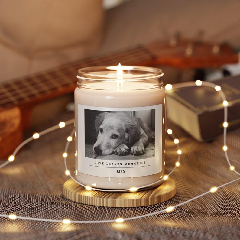 May include: A lit candle in a glass jar with a black and white photo of a dog on the front. The text "LOVE LEAVES MEMORIES" and the name "MAX" are below the photo. The candle sits on a round wooden coaster, with string lights around it.