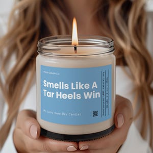 May include: A white candle with a blue label that reads "#Viral Candle Co. Smells Like A Tar Heels Win My Lucky Game Day Candle!" The candle is hand-poured with 100% natural soy wax and has a burn time of 50-60 hours.