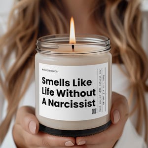 Puede incluir: Una vela beige con una etiqueta negra que dice "#Viral Candle Co. Smells Like Life Without A Narcissist" y un código QR. La vela está hecha a mano con cera de soja 100% natural y tiene un tiempo de combustión de 50 a 60 horas.