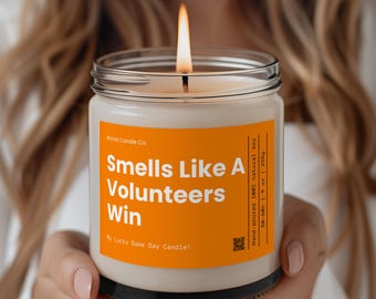 May include: A white candle in a clear glass jar with a black lid. The label is orange and says "Smells Like A Volunteers Win My Lucky Game Day Candle!"  Hand-poured 100% natural soy 50-60h 19 oz | 255g