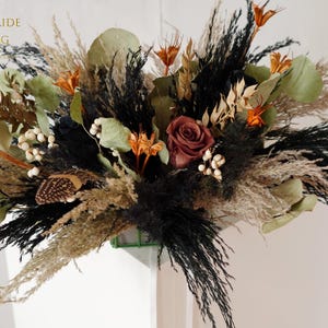 Puede incluir: Arreglo floral seco con una rosa burdeos, elementos negros, beige y naranja. El arreglo incluye hojas verdes y bayas blancas. El texto "PERFECTBRIDE WEDDING" es visible. Mezcla de texturas.