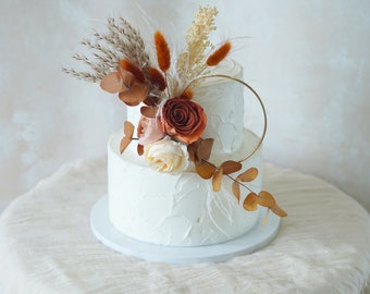 May include: Two-tiered white wedding cake decorated with dried flowers and greenery. The cake is topped with a gold ring and features a rustic, bohemian style.