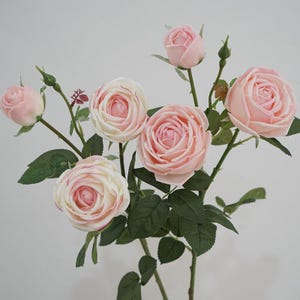 May include: A bouquet of artificial roses featuring soft pink and cream-coloured petals. The arrangement includes several fully bloomed flowers, smaller rosebuds, and dark green serrated leaves attached to thin, upright green stems.