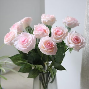 May include: A bouquet of artificial roses featuring soft pink petals that transition to a deeper rose-pink centre. The flowers are arranged on green stems with detailed green leaves, all displayed in a clear glass vase with visible stems.