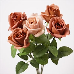May include: A bouquet of five artificial roses featuring four deep terracotta-coloured blooms and one central pale blush-pink rose. Each flower is attached to a green stem with detailed serrated leaves, creating a realistic floral arrangement.
