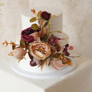 May include: A two-tiered white cake with horizontal ridges, decorated with an arrangement of artificial flowers in shades of deep burgundy, dusty pink, and muted orange, accented with small dark berries and green foliage.