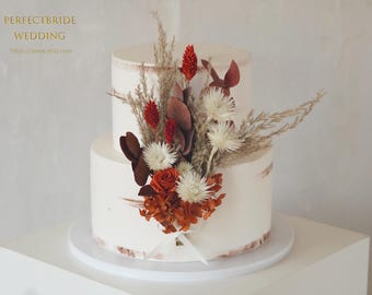 May include: Two-tiered white wedding cake decorated with dried flowers in shades of red, orange, and cream. The cake has a rustic design with visible brushstrokes of frosting. The text "PERFECTBRIDE WEDDING" is visible in the upper left corner.