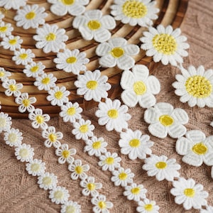 May include: Close-up of white daisy flower trim with yellow centers. The trim is arranged in rows and is made of a delicate, embroidered fabric. The daisies are placed on a brown woven surface.